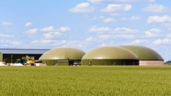 General view of a biogas plant with three digesters in a green wheat field in the countryside under a blue sky with white clouds.